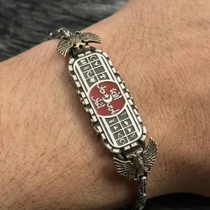 May include: A silver-toned metal bracelet featuring a central rectangular plaque with a red circular inlay containing white Arabic script and a crescent moon. The plaque is flanked by two winged motifs and connected to a chain with a textured link design.