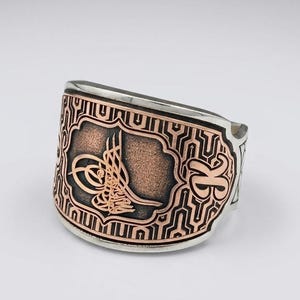 Peut inclure: Une large bague en argent avec un panneau central rectangulaire couleur cuivre orné d'un emblème calligraphique tughra. La surface environnante présente un motif géométrique noir en labyrinthe et la lettre K est gravée sur le côté droit.