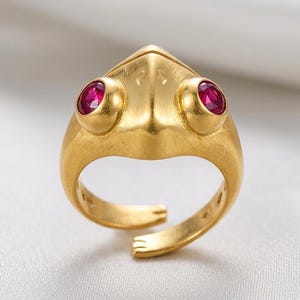 May include: A gold-toned ring shaped like a frog head with a brushed matte finish. The ring features two prominent, round, vibrant red gemstones set as eyes on either side of the top. The band is open at the bottom, showing a small foot-like detail.