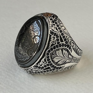Puede incluir: Un anillo de plata con una piedra ovalada negra con caligrafía árabe. La banda está decorada con intrincados diseños florales y piedras preciosas negras.
