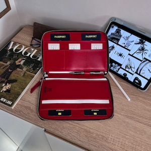 May include: Open red leather travel wallet with multiple compartments for passports and cards. The wallet is open, revealing the interior with a pen and documents. A magazine and tablet are in the background.