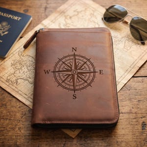 Puede incluir: Un organizador de documentos de viaje de cuero marrón con un diseño de rosa de los vientos grabado en oscuro en la portada. El estuche rectangular tiene una cremallera lateral y está colocado sobre un mapa antiguo con un pasaporte y gafas de sol.