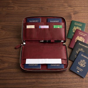 Könnte beinhalten: Ein dunkelroter Reiseorganizer aus Leder mit Reißverschluss, der über mehrere Innenfächer für vier blaue Reisepässe, mehrere Kreditkarten und einen schwarzen Stift verfügt. Vier weitere Reisepässe in Grün, Kastanienbraun, Schwarz und Blau liegen daneben.