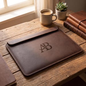 Könnte beinhalten: Eine dunkelbraune Laptoptasche aus Leder mit Klappverschluss und einer geprägten Monogramm-Gravur der Buchstaben AB. Die Tasche hat sichtbare Nähte an den Kanten und liegt auf einer Holzoberfläche neben einer Keramiktasse und Büchern.