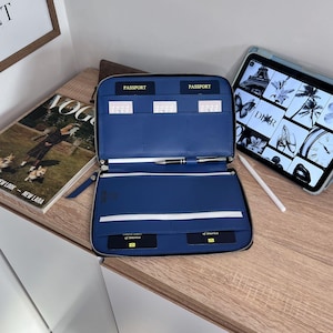 Op de afbeelding: Open blauw leren reisportemonnee met meerdere compartimenten voor paspoorten, kaarten en een pen. De portemonnee is open en toont de binnenkant. Een tijdschrift en een tablet met stylus zijn zichtbaar op de achtergrond.