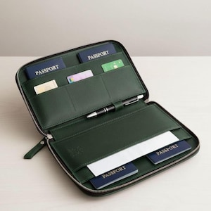 May include: A dark green zippered travel organizer featuring multiple interior slots for documents and cards. The organizer contains two blue passports labeled PASSPORT, a black pen, and several white and colored cards tucked into the various pockets.