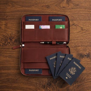 May include: A brown leather travel organizer with a zipper closure, featuring multiple interior slots for passports and credit cards, a central pen loop with a black pen, and four dark blue United States passports arranged on the exterior.