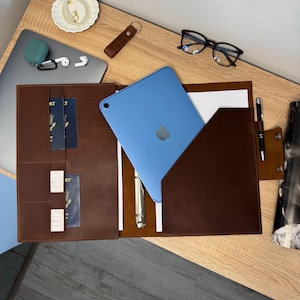 May include: A brown leather folio with a light blue tablet and a pen. The folio is open, revealing pockets for cards and passports. A silver pen and a blue tablet with the Apple logo are visible. Other items include glasses, a keychain, and a laptop.