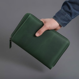 May include: A dark green rectangular zipped folio case with a smooth matte finish. The case features a matching green zip closure with a small rectangular pull tab. The design is simple and functional, providing a secure storage solution.