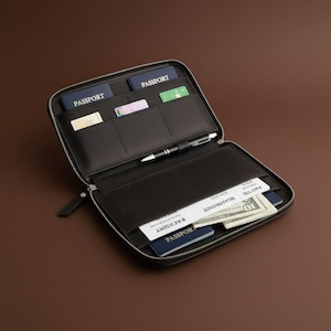 May include: Open black leather travel wallet with multiple compartments. The wallet contains passports, credit cards, cash, and a pen. The wallet is zippered and has a small handle.