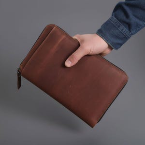 May include: A brown leather wallet with a zipper closure. The wallet is rectangular and appears to be made of high-quality leather. The zipper runs along three sides of the wallet. The wallet is held by a person.