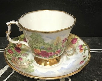 May include: A white teacup and saucer with a floral pattern featuring roses, a landscape scene, and gold trim.