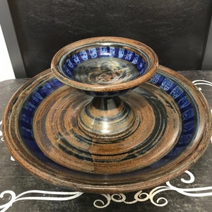 Può includere: Un piatto da portata in ceramica a due livelli con una glassa marrone e blu. Il livello superiore è una piccola ciotola e il livello inferiore è un piatto più grande. Il piatto ha un aspetto rustico e fatto a mano.