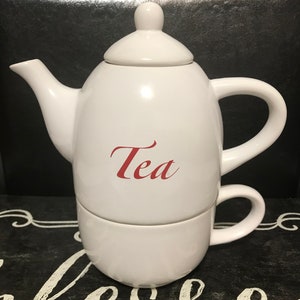 Könnte beinhalten: Weiße Teekanne aus Keramik mit Deckel und passender Tasse. Die Teekanne hat das Wort "Tea" in rot auf der Seite.