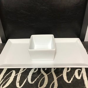 May include: A white rectangular serving platter with a square bowl in the centre. The platter and bowl are ceramic. The background is a black chair.