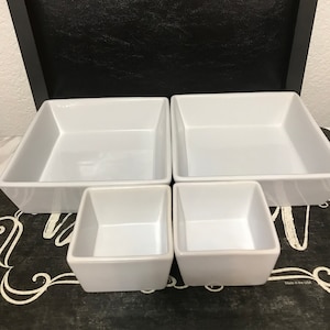 Op de afbeelding: Set van vier witte keramische serveerschalen. De schalen zijn vierkant en in verschillende maten. De grootste schaal is achteraan, de kleinste schaal vooraan. De schalen staan op een zwart oppervlak met witte letters.