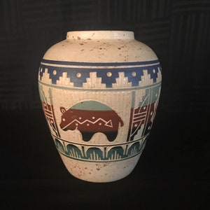 May include: A white ceramic vase with a southwestern design. The vase features a brown, green, and blue geometric pattern with a stylized animal figure.