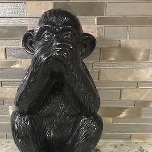 Puede incluir: Una figurita de mono negro sentado con las manos sobre los ojos. El mono está hecho de un material liso y brillante y tiene una apariencia realista.