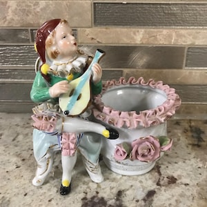 May include: A ceramic figurine of a jester playing a lute, seated next to a small white planter with pink trim and roses. The jester wears a green jacket, red hat, and light blue pants. The planter is approximately 4 inches tall.