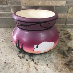May include: A hand-painted ceramic pot with a wide opening and a rounded body. The pot is painted in shades of purple and features a white bison, a sunset scene, and black tree silhouettes.