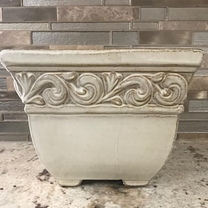 May include: A white ceramic planter with an ornate design around the top edge. The planter has a square shape with rounded corners and sits on four small feet.