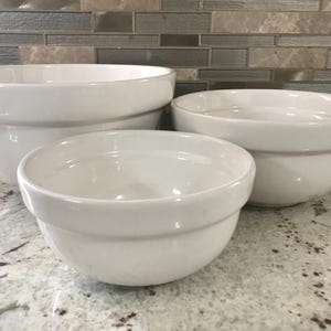 May include: Three white ceramic mixing bowls of different sizes. The bowls are nested inside each other.