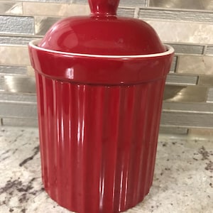 Pode incluir: Um pote de cerâmica vermelho com tampa branca e lados canelados. O pote é perfeito para guardar açúcar, café, chá ou outros produtos secos.