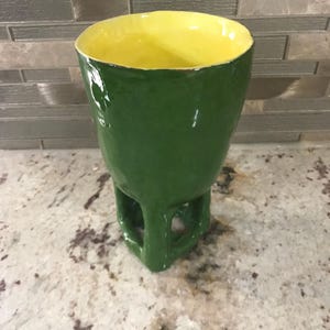 Può includere: Tazza in ceramica fatta a mano con esterno verde vibrante e interno giallo brillante. La tazza ha un'esclusiva base a forma di piedistallo con dettagli ritagliati. La finitura lucida esalta i colori, rendendola un pezzo d'effetto.