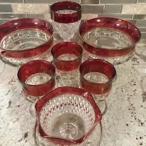 Può includere: Una collezione di piatti e bicchieri di vetro trasparente con bordi rosso rubino. Il set include ciotole, bicchieri piccoli e un bicchiere alto, tutti con un motivo a taglio diamante. I bordi rosso rubino aggiungono un tocco di colore.