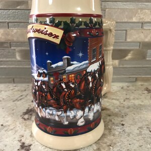 Può includere: Un boccale di birra in ceramica con la parte superiore e la base color crema. Il corpo presenta una scena invernale con una squadra di cavalli che traina una carrozza davanti a un edificio. La parola "Budweiser" è visualizzata su uno striscione.
