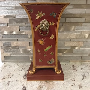 Könnte beinhalten: Eine dekorative Vase mit burgunderfarbenem Hintergrund und goldenem Rand. Die Vase hat einen Löwenkopf-Ringgriff und Insektenillustrationen. Der Sockel hat goldfarbene Füße.