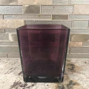 May include: A square, dark purple glass vase with a thick base. The vase has a slightly tapered shape and is empty. The background features a neutral-toned, textured tile backsplash.