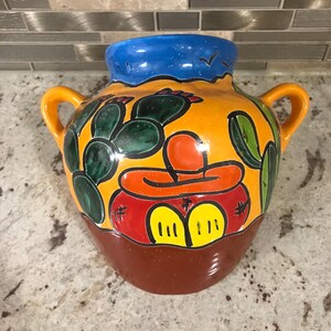 Könnte beinhalten: Eine handbemalte Keramikvase mit gelbem Hintergrund und blauem Rand. Die Vase zeigt ein Kaktus-Design mit grünen, roten und gelben Akzenten. Die Vase hat zwei Henkel.