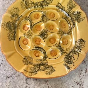 May include: Yellow ceramic escargot plate with 12 individual wells. The plate has a scalloped edge and is decorated with black and green leaf-like patterns. Each well contains a brown circle.