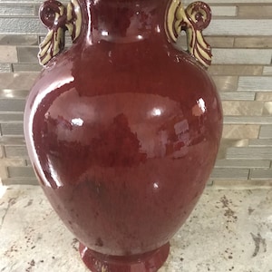 May include: A large, glossy, burgundy-colored ceramic vase with two ornate handles. The vase has a wide, rounded body and a slightly flared base. The handles are a lighter shade of beige and brown.