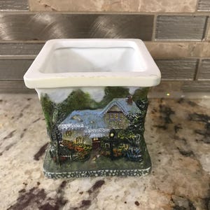 May include: A white ceramic planter with a square top and a detailed, three-dimensional depiction of a cottage scene. The cottage has a blue roof, surrounded by green trees and foliage. The base is a stone-like texture.