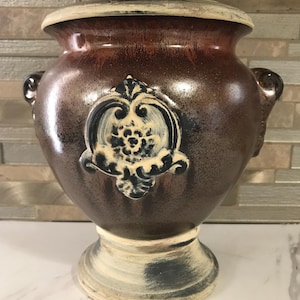 May include: A brown ceramic vase with a white and black design. The vase has a wide base and a narrow neck. It has two handles on the sides.