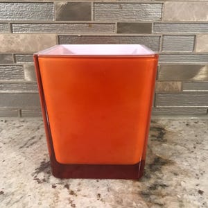 May include: A square orange glass vase with a white interior. The vase has a dark red base. The vase is empty and ready to be filled with flowers or other decorative items. The background is a neutral tone.