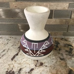 Pode incluir: Um pequeno vaso de cerâmica branco sujo com uma base bulbosa e um topo alargado. A base apresenta um design geométrico em tons de castanho, roxo e branco. O vaso tem um pescoço estreito e uma abertura larga.