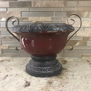 May include: A decorative urn with a red body and a black base. The urn has two curved handles and a detailed rim. The base is also black with a decorative pattern. The urn is likely made of metal or a similar material.