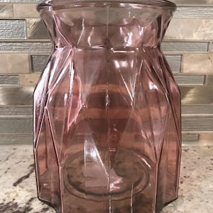 Puede incluir: Un jarrón de vidrio transparente con un patrón geométrico. El jarrón es de color rosa oscuro y tiene una boca ancha.