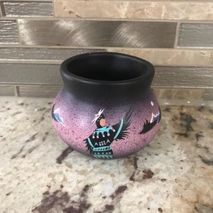 May include: A small, round, hand-painted ceramic pot with a black interior and a pink and purple exterior. The pot features a Native American figure with turquoise accents and a mountain scene. The pot is sitting on a speckled surface.