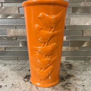 May include: Orange ceramic vase with a design of five birds in relief. The birds are arranged in a vertical stack, with the largest bird at the top and the smallest bird at the bottom.