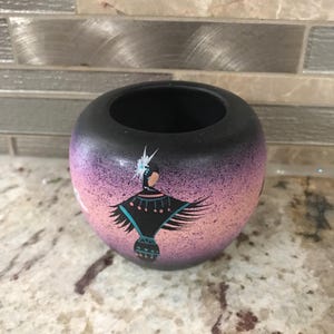 May include: A round, black ceramic pot with a Native American design. The pot features a gradient of pink and purple with a painted figure in traditional attire. The pot is sitting on a speckled countertop.