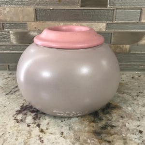 May include: A round, gray ceramic jar with a pink lid. The jar has a smooth, matte finish and a small, circular opening at the top. The lid is a lighter shade of pink and sits snugly on the jar.