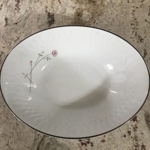 May include: White oval serving bowl with a delicate pink rose and stem design. The bowl has a textured surface and a thin black trim around the edge. A classic piece for serving salads or sides.