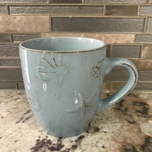 Może przedstawiać: Jasnoniebieski ceramiczny kubek z wytłoczonymi wzorami muszli i rozgwiazd. Kubek ma brązową obręcz i uchwyt, odpowiedni do gorących lub zimnych napojów. Motyw nadmorski.