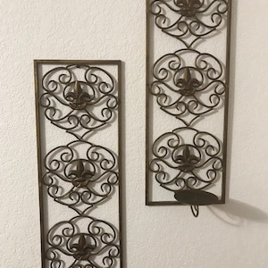 Puede incluir: Dos apliques de pared decorativos de metal con un diseño de flor de lis. Cada aplique tiene tres motivos de flor de lis y un portavelas.