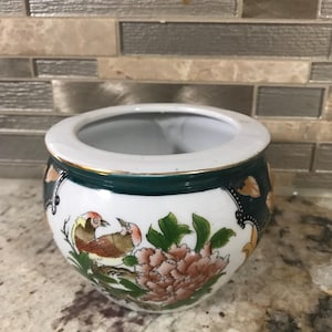 Könnte beinhalten: Eine kleine, runde, weiße Keramikschale mit einem grünen Band und goldenem Rand. Die Schale hat ein gemaltes Design von zwei Vögeln, rosa Blumen und grünen Blättern. Die Schale ist wahrscheinlich für dekorative Zwecke.