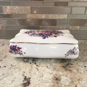 May include: A white ceramic trinket box with a floral design on the lid and sides. The lid features a bouquet of purple grapes and pink, purple, and yellow flowers. The box has a gold trim.